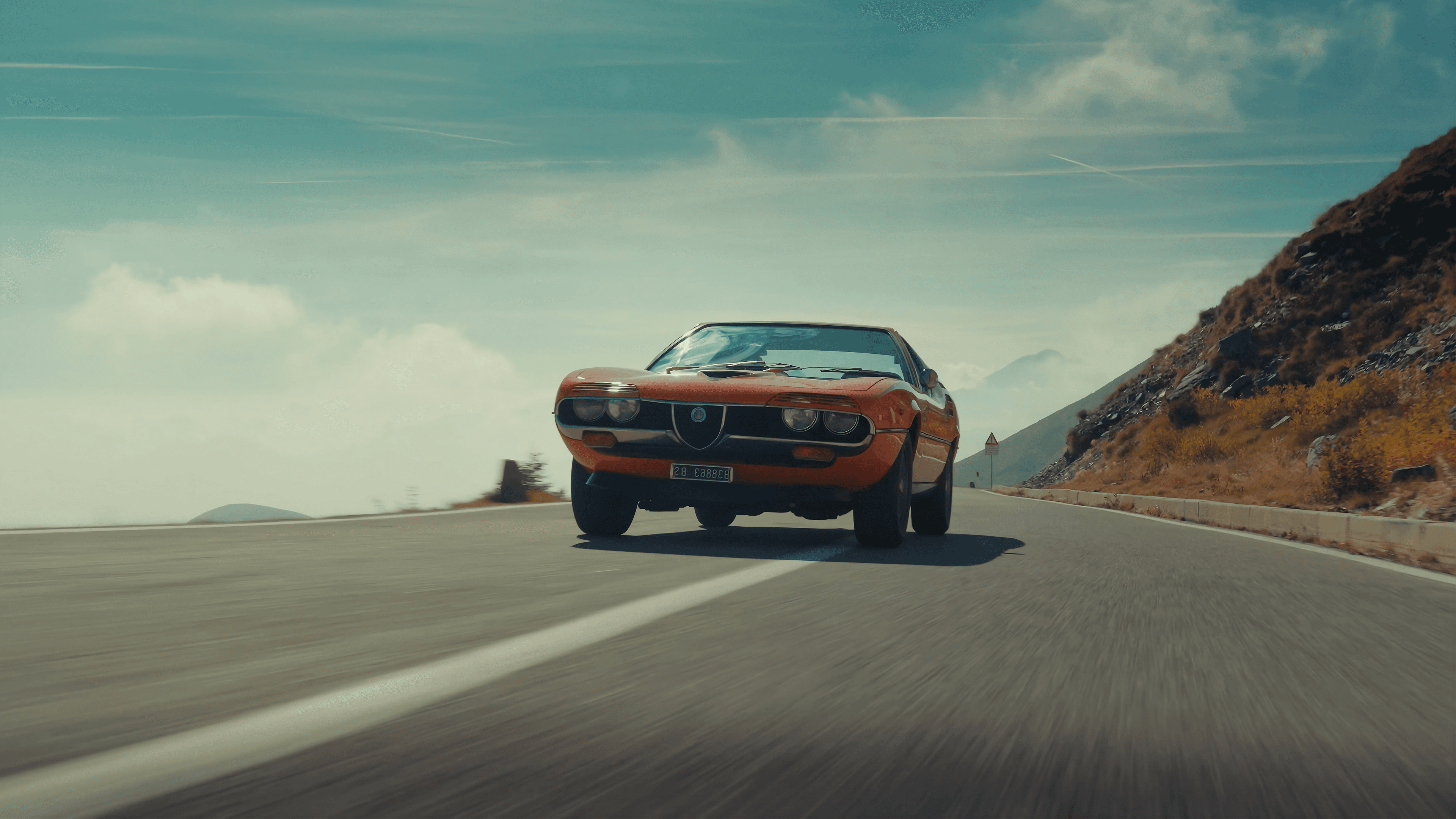 Alfa Romeo Montreal arancione che percorre una strada di montagna sotto un cielo azzurro.